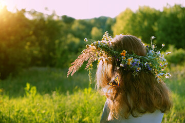 Girl in flower wreath close up on sunny meadow, rear view. Floral crown, symbol of summer solstice....