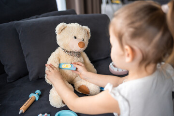 Little girl playing doctor with toys and teddy bear on the sofa in living room at home