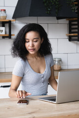 Portrait of attractive African American woman sitting at the kitchen table with a laptop and using a mobile phone, answering an online call, typing a message. The girl is busy with gadgets