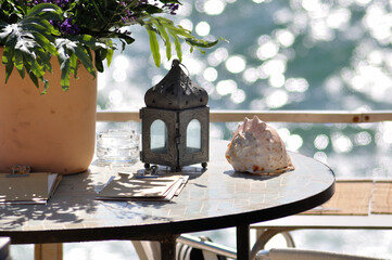 A lamp, whelk vase with plant, and a notebook on top of a table near the sea