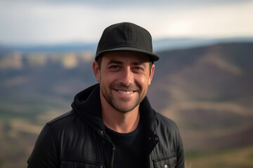 Portrait of a handsome young man in a black leather jacket and cap smiling at the camera on the top of a mountain