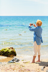 Elderly woman standing on the beach by the sea and taking photo of the sea and seagulls on mobile phone. Active seniors concept
