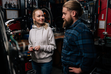 Fototapeta na wymiar Bearded repairman male communicating with smiling blonde female client, talking problem of bicycle, detected during diagnostics in repair shop with dark interior, standing by MTB.