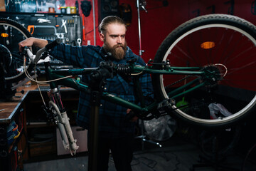Fototapeta na wymiar Concentrated bearded repairman watching rear shifter of mountain bike, changing speeds using handlebar shift lever working in bicycle repair shop with dark interior. Concept of maintenance of bicycle.