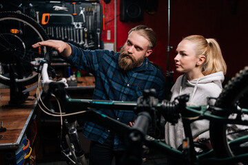 Fototapeta na wymiar Bearded cycling repairman communicating with blonde female client, talking about problem of bicycle detected during diagnostics in repair shop with dark interior. Concept of bike maintenance.