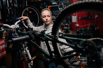 Focused cycling mechanic female watching rear shifter of mountain bike, changing speeds using handlebar shift lever, working in bike repair workshop with dark interior. Concept of bike maintenance. - Powered by Adobe
