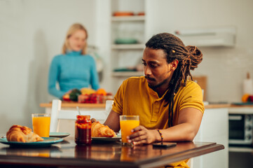 A multicultural man is having breakfast at home while his wife is standing in a kitchen in a blurry background.