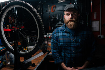 Portrait of bearded cycling mechanic male in cap posing standing by bicycle in bike repair workshop with dark interior, looking at camera with serious expression. Concept of bicycle maintenance.
