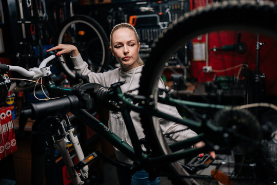 Focused cycling mechanic female watching rear shifter of mountain bike, changing speeds using handlebar shift lever, working in bike repair workshop with dark interior. Concept of bike maintenance.