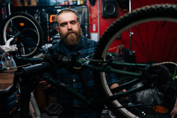 Portrait of handsome bearded cycling repairman with fashion haircut standing by bicycle in repair workshop with dark interior, looking at camera. Concept of professional maintenance of bicycle.