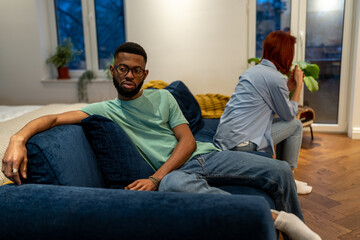 Unhappy young interracial couple sit separately on sofa at home, tired of each other, upset family man and woman not talking after fight, sit on couch apart. Bad communication in relationship