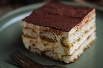 A piece of Tiramisu cake, traditional Italian dessert, served in moody, rustic scene. Wooden background and green plate. 