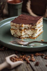 A piece of Tiramisu cake, traditional Italian dessert, served in moody, rustic scene. Wooden background and green plate. 