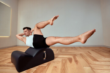 Fototapeta premium A young man does Pilates exercises on a reformer to strengthen the pelvic region of the musculoskeletal system, Pilates exercises on reformers. copy space
