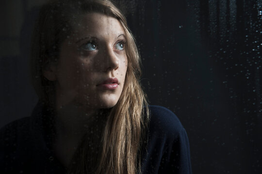 Portrait of young woman behind window, wet with raindrops, looking up