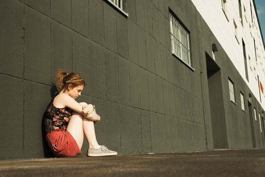 Teenage girl sitting on ground and leaning against wall, looking downwards