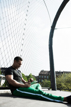 Mature man sitting on outdoor basketball court, looking at tablet computer, Germany