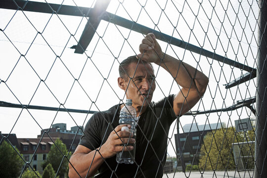 Mature man leaning against chain-link fence on outdoor basketball court, holding bottle of water, Germany