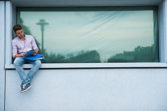 Young man sitting on ledge outdoors, looking at tablet computer, Germany