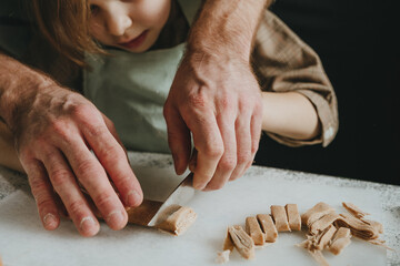 Unrecognizable father and daughter in aprons cut the dough on a white background against a dark wall