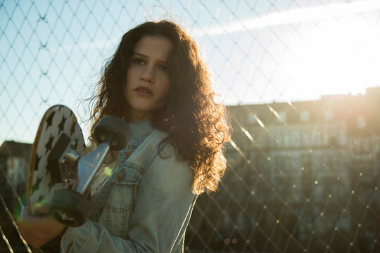 Portrait of teenage girl standing outdoors next to chain link fence, holding skateboard, Germany