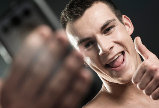 Close-up portrait of young man taking a selfie with cell phone, giving the thumbs up sign, studio shot on black background