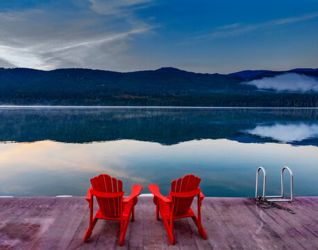 Two red Adirondack chairs on a dock with ladder on Sproat Lake at sunset, Vancouver Island, BC, Canada; Vancouver Island, British Columbia, Canada