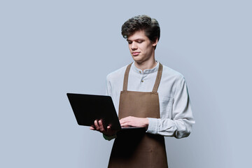 Young male in an apron with laptop, on gray studio background