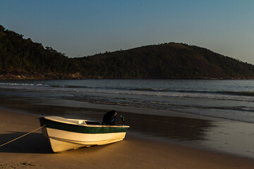 Barquinho branco de frente aportado na areia da praia, iluminado pela luz do sol de um final de tarde, com mar e montanhas ao fundo.