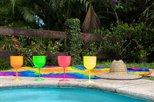 beira de piscina com copos coloridos de drink, canga colorida e chap&eacute;u de palha, com cerca de madeira e vegeta&ccedil;&atilde;o ao fundo