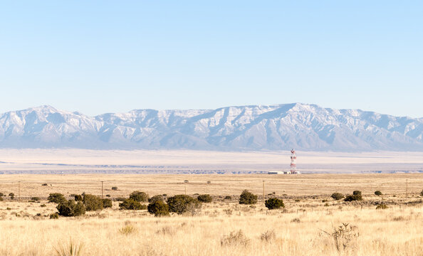 Snow-dusted Sandia Mountains from a distance. Middle distance a cell tower and building. Foreground the New Mexico High Plains. Full sun.