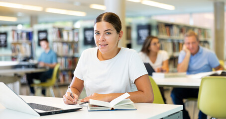 Concentrated young woman checking planner while studying in the library and using laptop