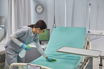 Young nurse preparing bed for patient cleaning it with sanitizer and rag in hospital ward - obrazy, fototapety, plakaty