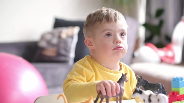 Cute litle boy living with down syndrome playing at home with animal toys holding it in hand and showing funny facial expressions, exploring new games