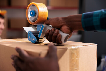 African american storehouse employee arms wrapping cardboard package with sticky tape. Post office worker hands packing customer order and closing parcel using adhesive dispenser closeup