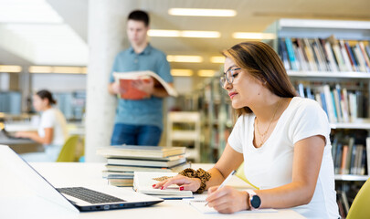 Fototapeta premium Concentrated young woman in glasses checking planner while studying and using laptop in the library