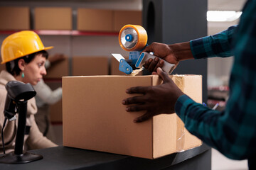 Factory warehouse worker packing freight for transportation using adhesive tape gun. African...