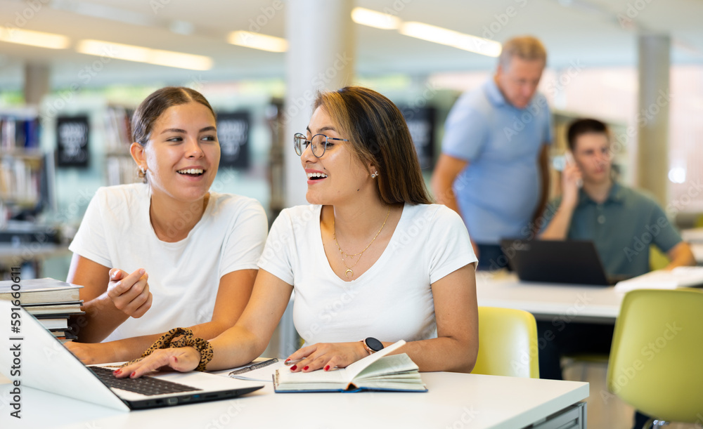 Poster two positive ladies wearing casual clothes behave loudly while studying in the library and using com
