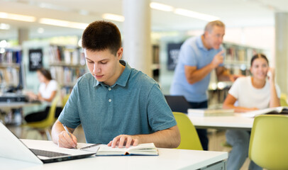 Concentrated young man checking plans in calendar while studying in the library and using laptop