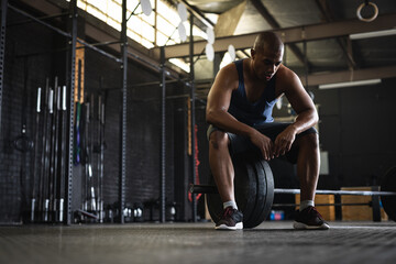 Full length of tired biracial young man with shaved head sitting on barbell in gym, copy space