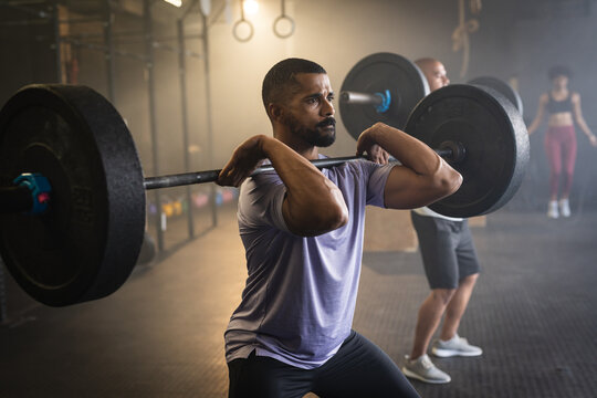 Dedicated biracial bearded young man lifting barbell while standing in gym - Powered by Adobe