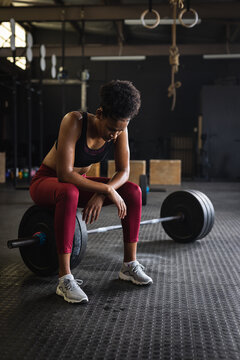 Full length of tired biracial young woman with afro hair sitting on barbell in gym, copy space