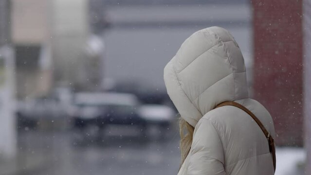 A woman in a hooded parka watches cars drive by as the snow falls
