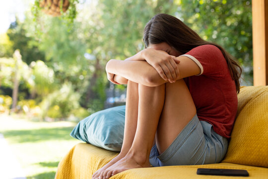 Sad caucasian teenager girl sitting on sofa, covering her face and crying
