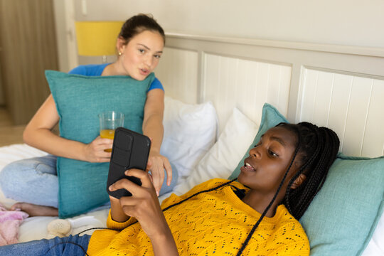 Happy diverse teenager girls friends sitting on bed, using smartphone and talking