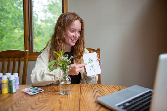 Smiling young woman holding a watercolor painting and explaining the art digitally on a computer in an online class from her home
