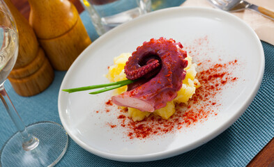 Appetizing baked octopus tentacle served on white plate with boiled potatoes garnished with paprika and greens..