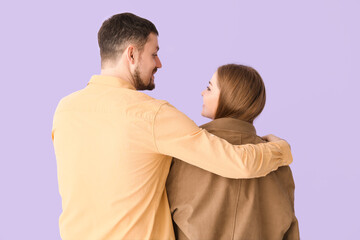 Young man hugging wife in his jacket on lilac background, back view