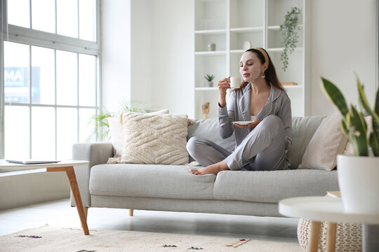 Young woman with sheet mask and cup of coffee sitting on sofa at home - Powered by Adobe