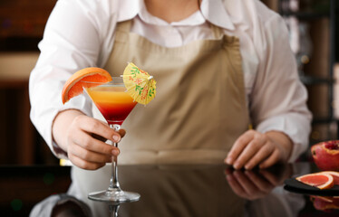 Female bartender holding glass of tasty Tequila Sunrise on dark table in bar